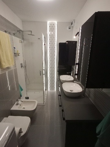 Nowoczesna łazienka z szarą glazurą, kabiną prysznicową ze szklanymi drzwiami, podwójną umywalką na czarnym blacie i toaletą typu bidet