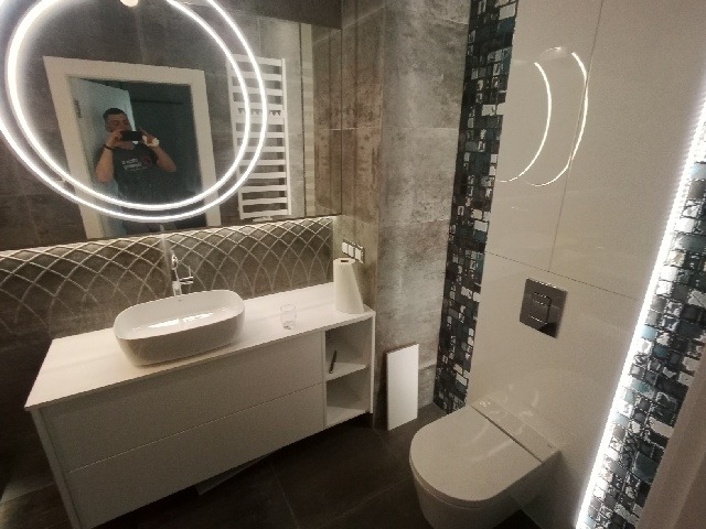 Nowoczesna łazienka z umywalką nablatową na białej szafce, okrągłym lustrem z podświetleniem LED, toaletą z ukrytym spłukiwaniem i dekoracyjnymi mozaikami na ścianach. W lustrze odbija się osoba...