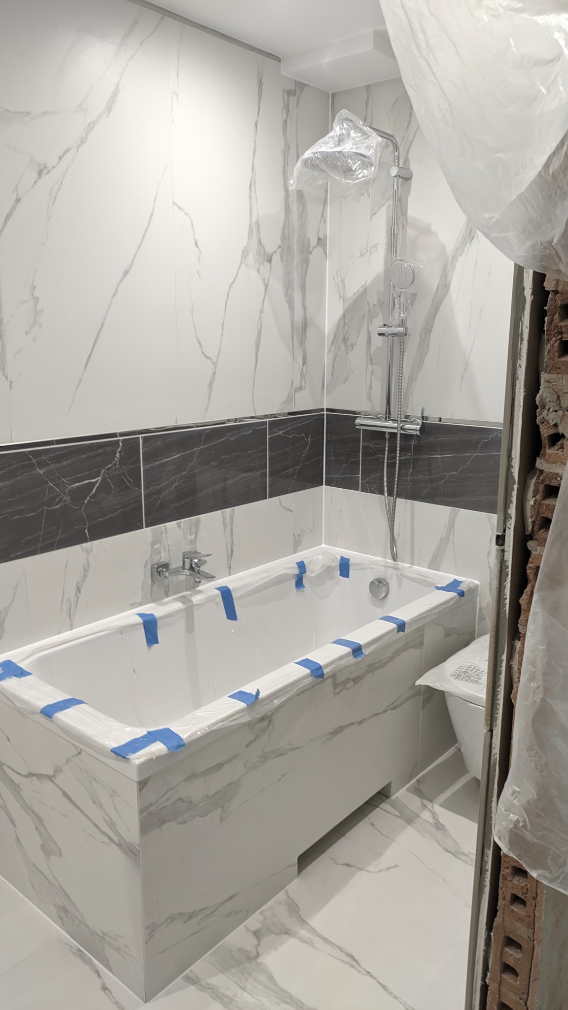 Łazienka w trakcie wykańczania: biała wanna z marmurkową obudową zabezpieczona niebieską taśmą, białe marmurkowe płytki na ścianach z pasem czarnych płytek, bateria wannowa i prysznic osłonięte...