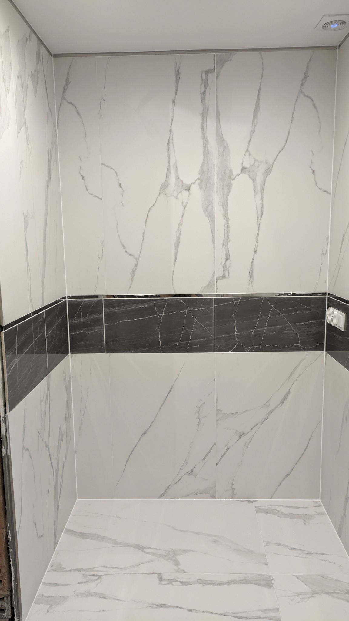 Wykończona kabina prysznicowa z białymi płytkami imitującymi marmur, z poziomym pasem czarnych płytek. Widoczny fragment podłogi również wyłożony płytkami w podobnym stylu, oraz gniazdko...