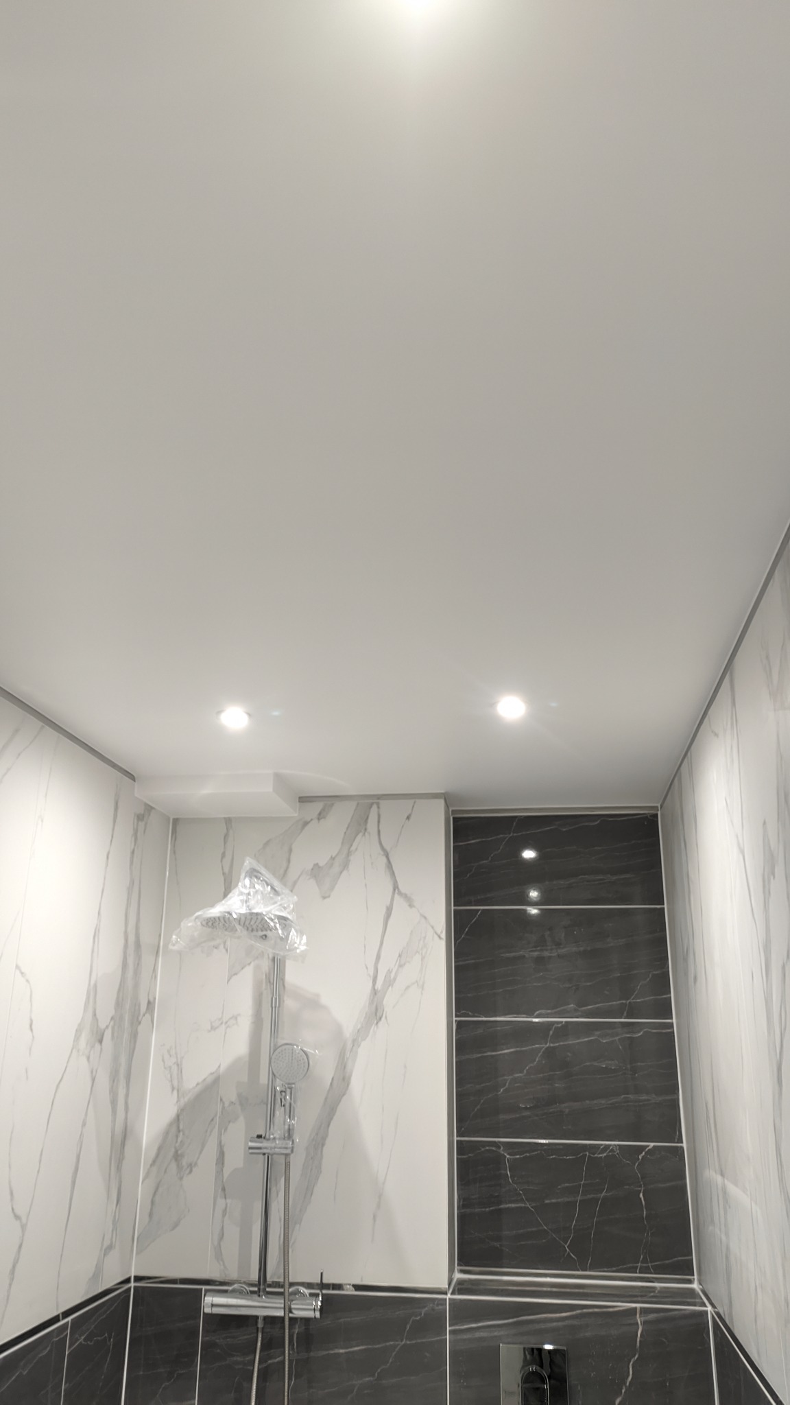 Nowoczesna kabina prysznicowa z chromowaną baterią i deszczownicą, częściowo jeszcze w folii ochronnej, otoczona białymi i czarnymi płytkami imitującymi marmur, podświetlona punktowymi lampami...