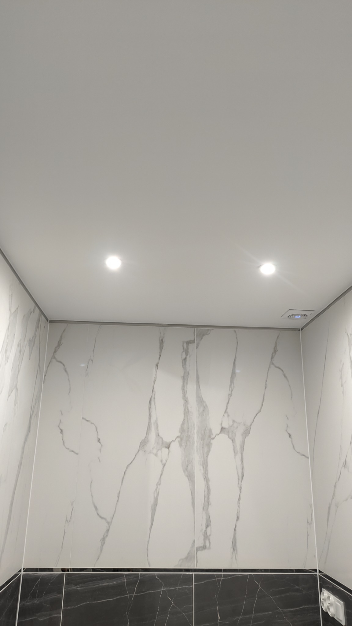 Wykończona łazienka z białymi płytkami imitującymi marmur na ścianach i czarnymi płytkami na dole, oświetlona dwoma punktowymi lampami sufitowymi i wentylatorem z niebieską diodą.