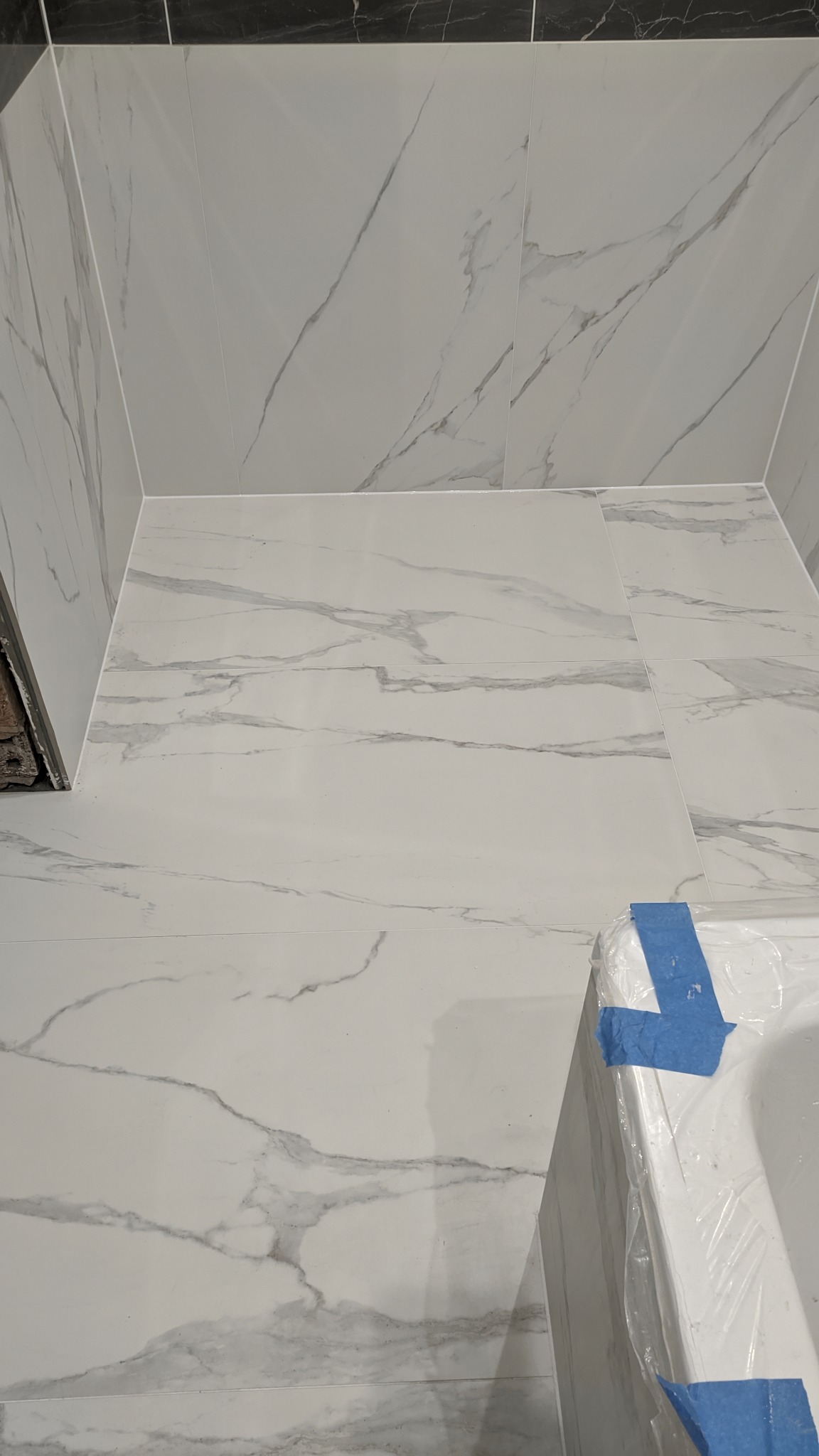 Świeżo wykafelkowana łazienka z białymi płytkami imitującymi marmur, zabezpieczona folią wanna z niebieską taśmą malarską w rogu pomieszczenia.