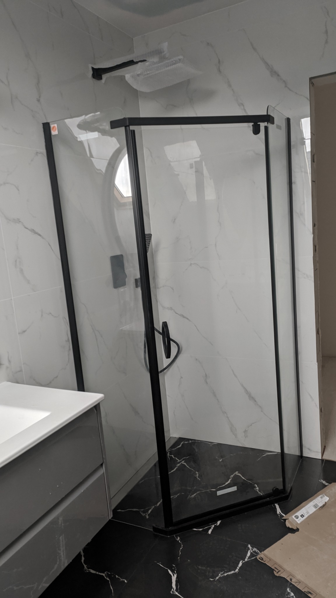 Nowoczesna kabina prysznicowa z czarnymi profilami na tle marmurowych płytek, widoczna umywalka i czarna podłoga z wzorem marmuru, element armatury w folii ochronnej.