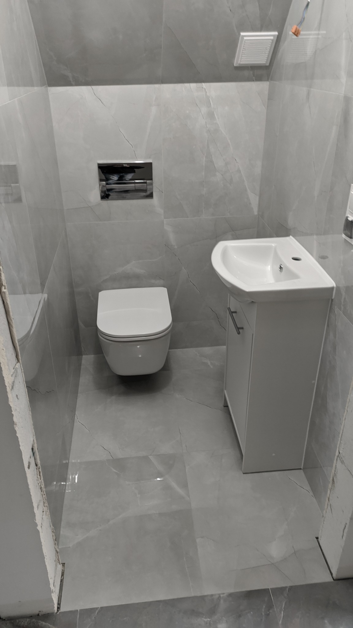 Wykończona łazienka z szarymi płytkami imitującymi marmur, wiszącą toaletą i białą szafką pod umywalką. Widoczny fragment niezakończonego obramowania drzwi.