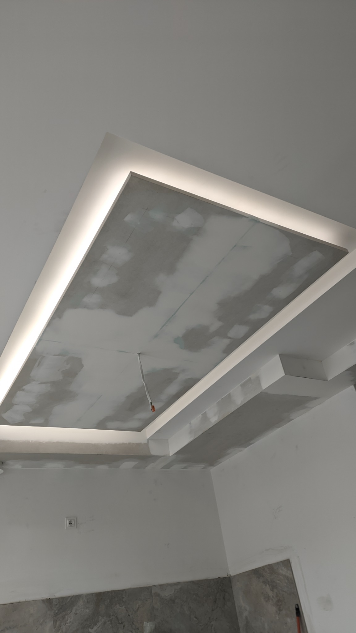 Sufit podwieszany z oświetleniem LED w trakcie szpachlowania, widoczne niedoskonałości i kabel wystający z sufitu, ściany w kolorze białym z fragmentem wykończenia imitującego marmur na dole.