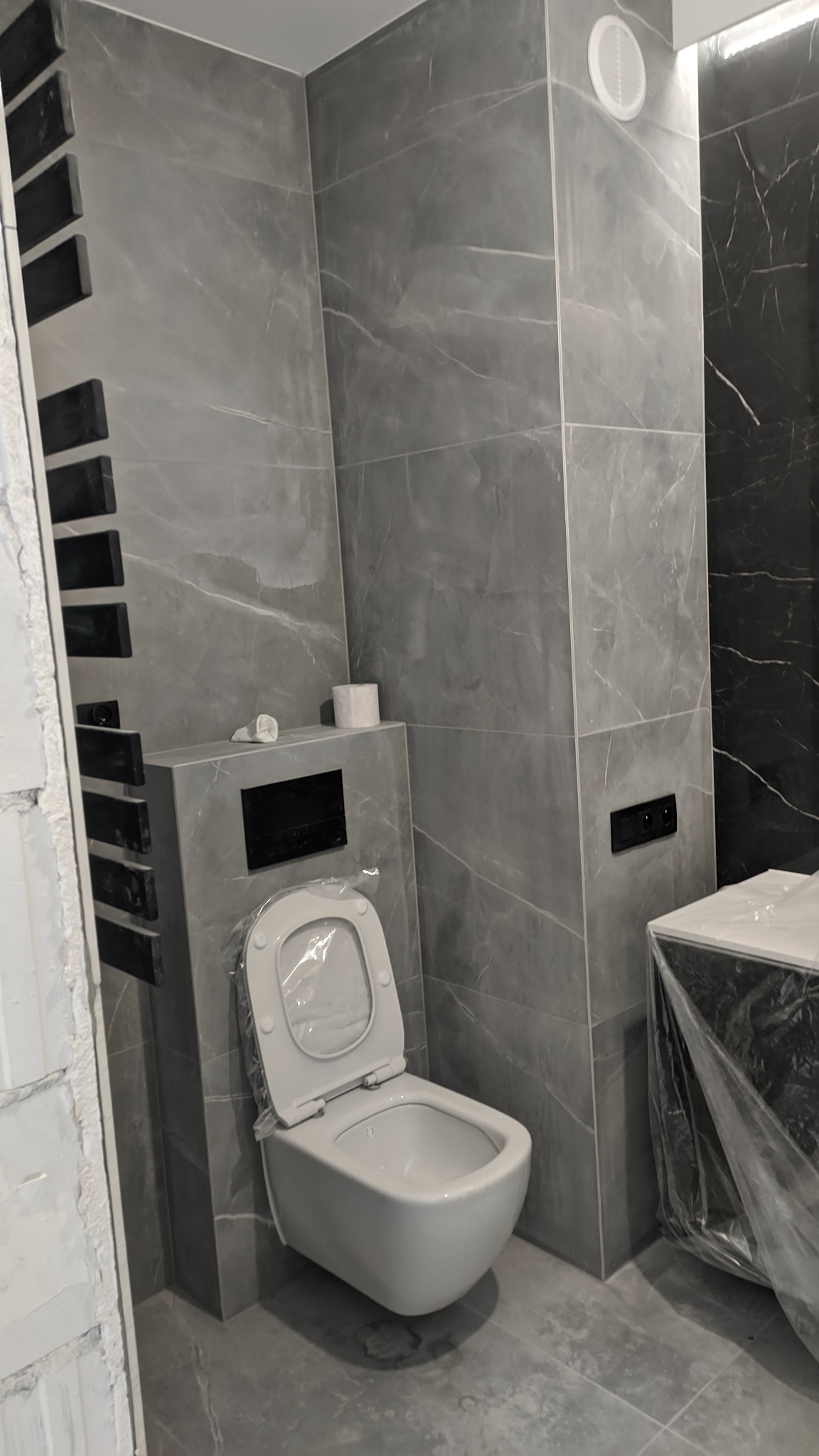 Nowoczesna toaleta podwieszana z szarą marmurkową okładziną ścienną, częściowo zakryta folią ochronną, widoczny fragment grzejnika drabinkowego i rolka papieru toaletowego.