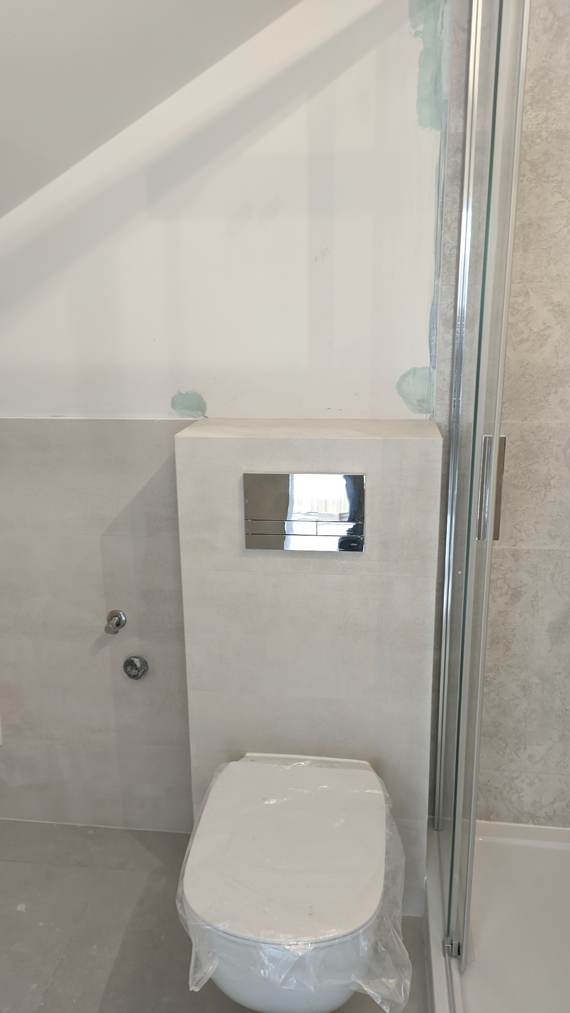 Nowa toaleta w folii ochronnej, zamontowana obok kabiny prysznicowej z chromowanymi detalami, widoczne ślady szpachli na ścianie nad zabudową stelaża podtynkowego.