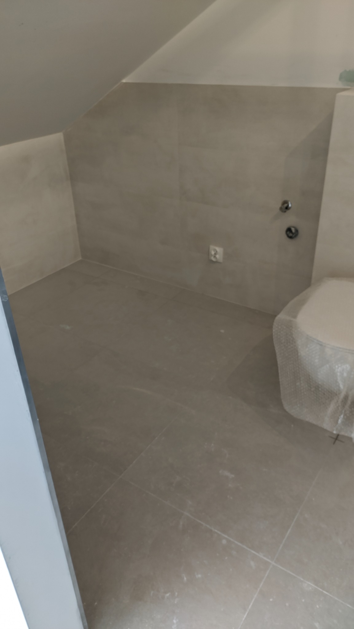 Wykończona łazienka na poddaszu z beżowymi płytkami podłogowymi i ściennymi, widoczne przyłącza wody i częściowo zakryta folią ochronną toaleta.