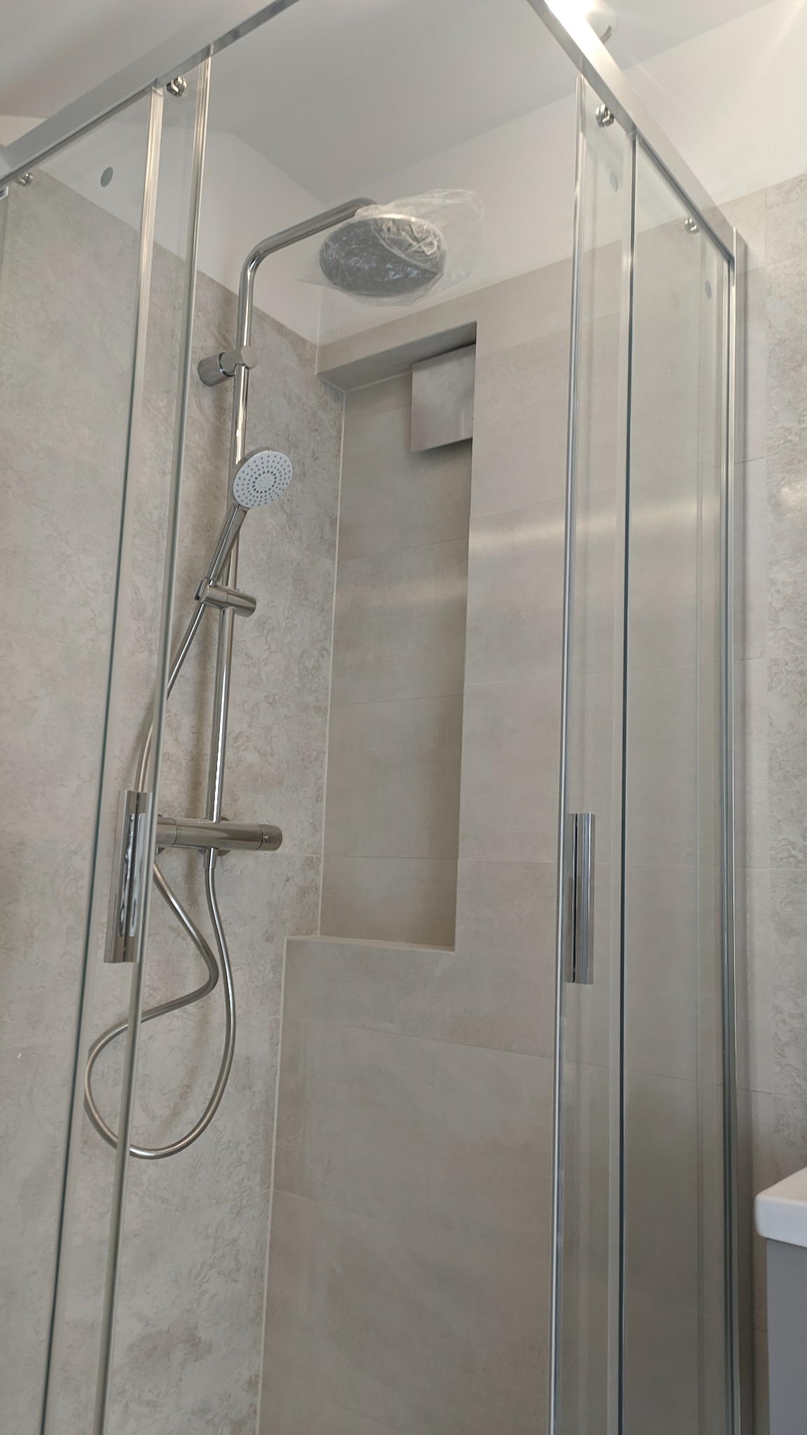 Nowoczesna kabina prysznicowa z deszczownicą w folii ochronnej, jasne płytki imitujące kamień, wnęka w ścianie wykończona tym samym materiałem.