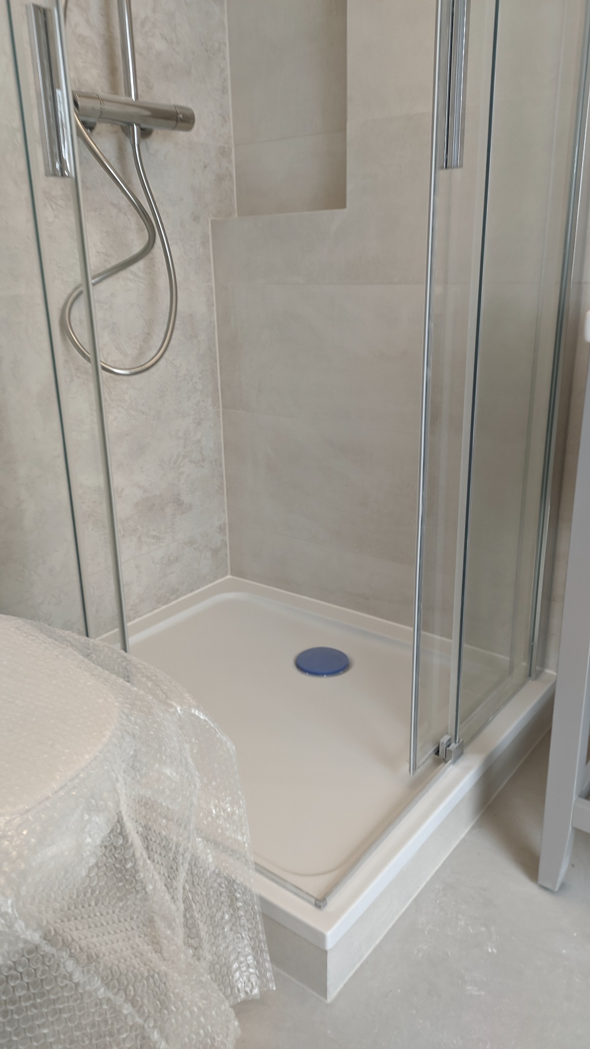 Nowy brodzik z kabiną prysznicową w trakcie montażu, zabezpieczony folią bąbelkową, z widoczną armaturą i szarymi płytkami na ścianie.