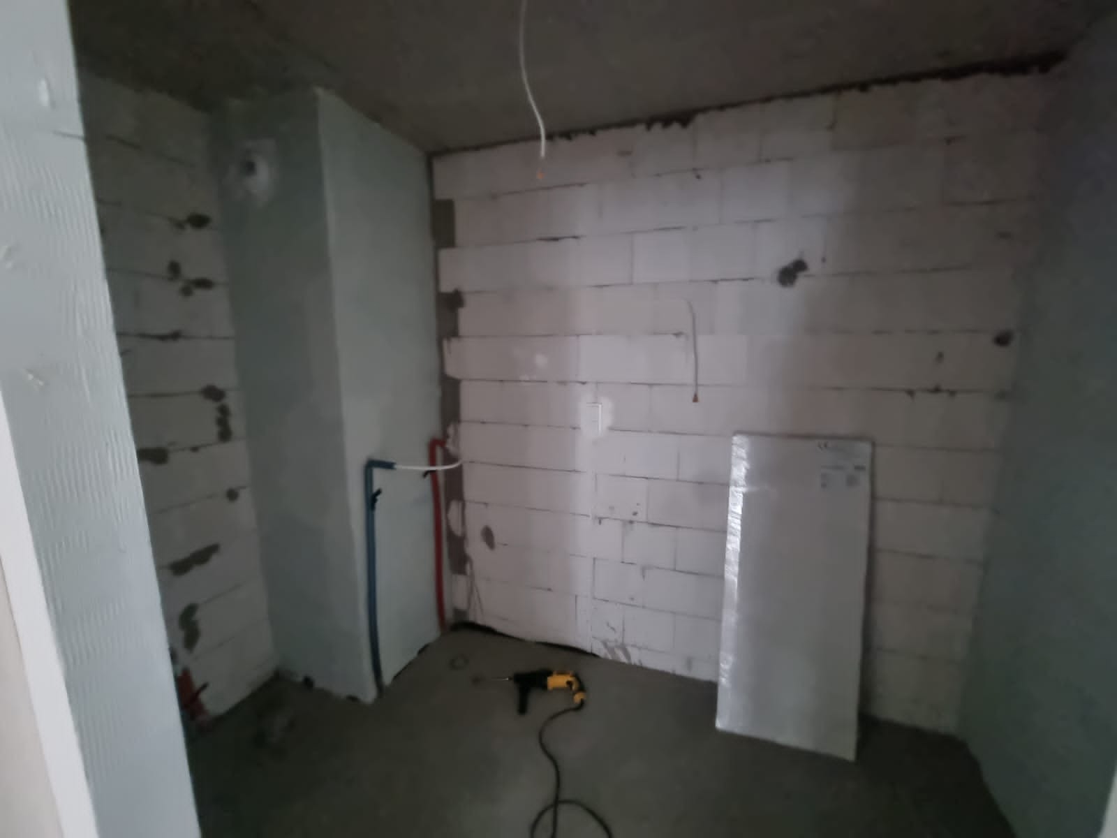 Surowy stan łazienki w trakcie prac: ściany z białych bloczków, widoczne instalacje wodne, przewód elektryczny zwisający z sufitu i wiertarka na podłodze.