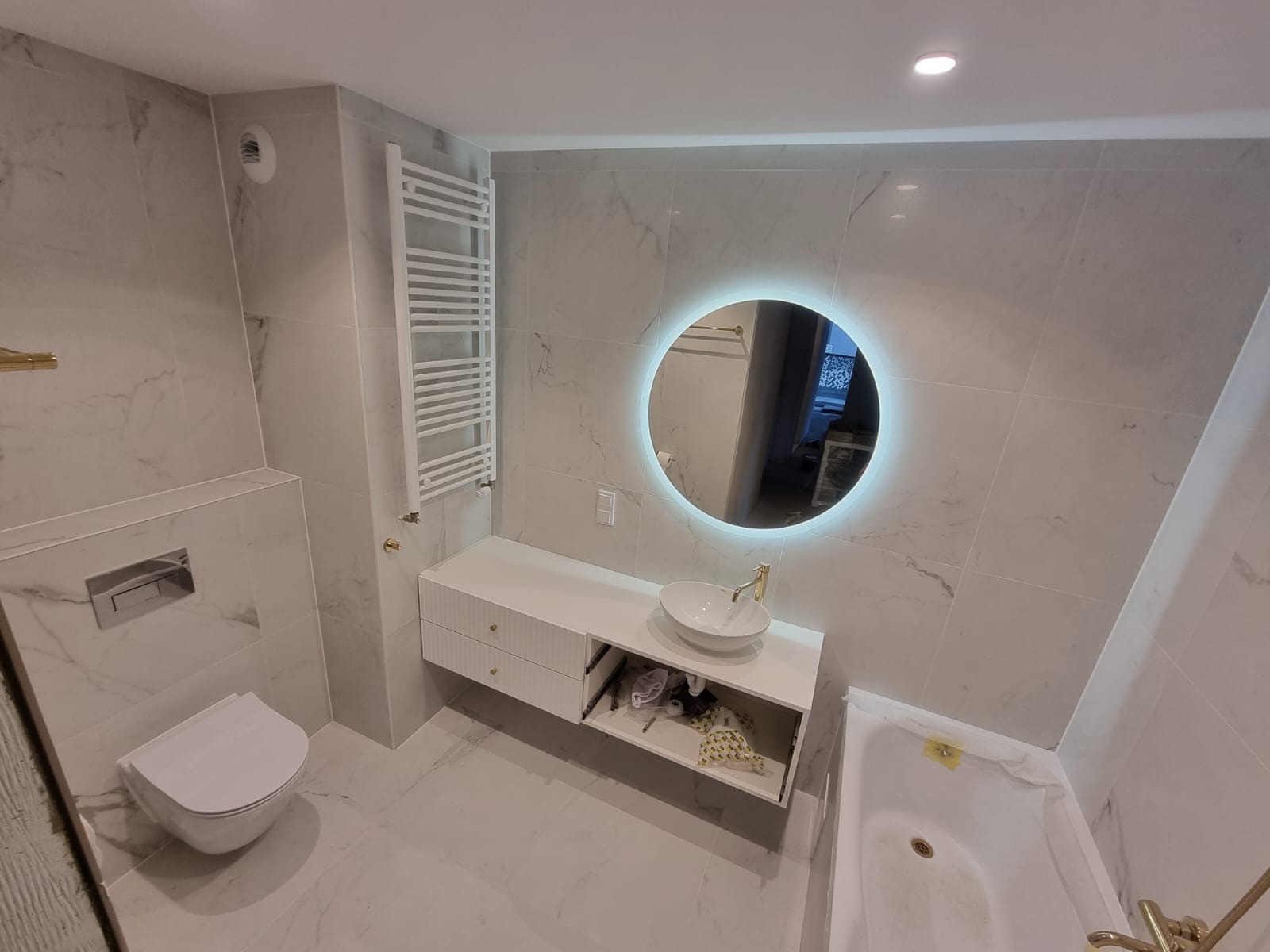 Nowoczesna łazienka wykończona białymi płytkami imitującymi marmur, z okrągłym lustrem podświetlanym LED, wiszącą szafką z umywalką i otwartą półką, grzejnikiem drabinkowym i podwieszaną toaletą.