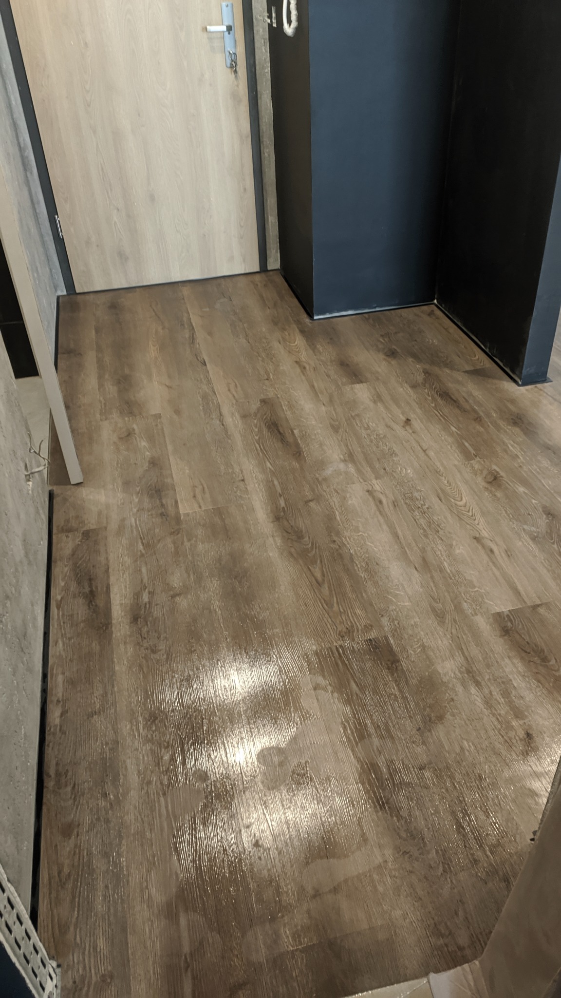 Nowo położona, błyszcząca podłoga laminowana w odcieniach brązu, w tle fragment drzwi wejściowych i czarnej ściany.