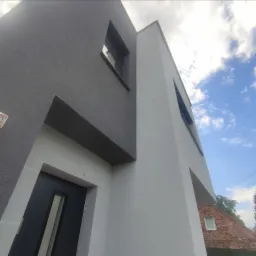 Dwukolorowa elewacja nowoczesnego domu z widoczną stolarką okienną i drzwiami wejściowymi, sfotografowana pod kątem z fragmentem nieba w tle.