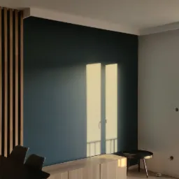 Nowoczesne wnętrze z ciemnozieloną ścianą, drewnianą ażurową ścianką działową, wiszącymi lampami i widokiem na osiedle przez balkonowe okno.