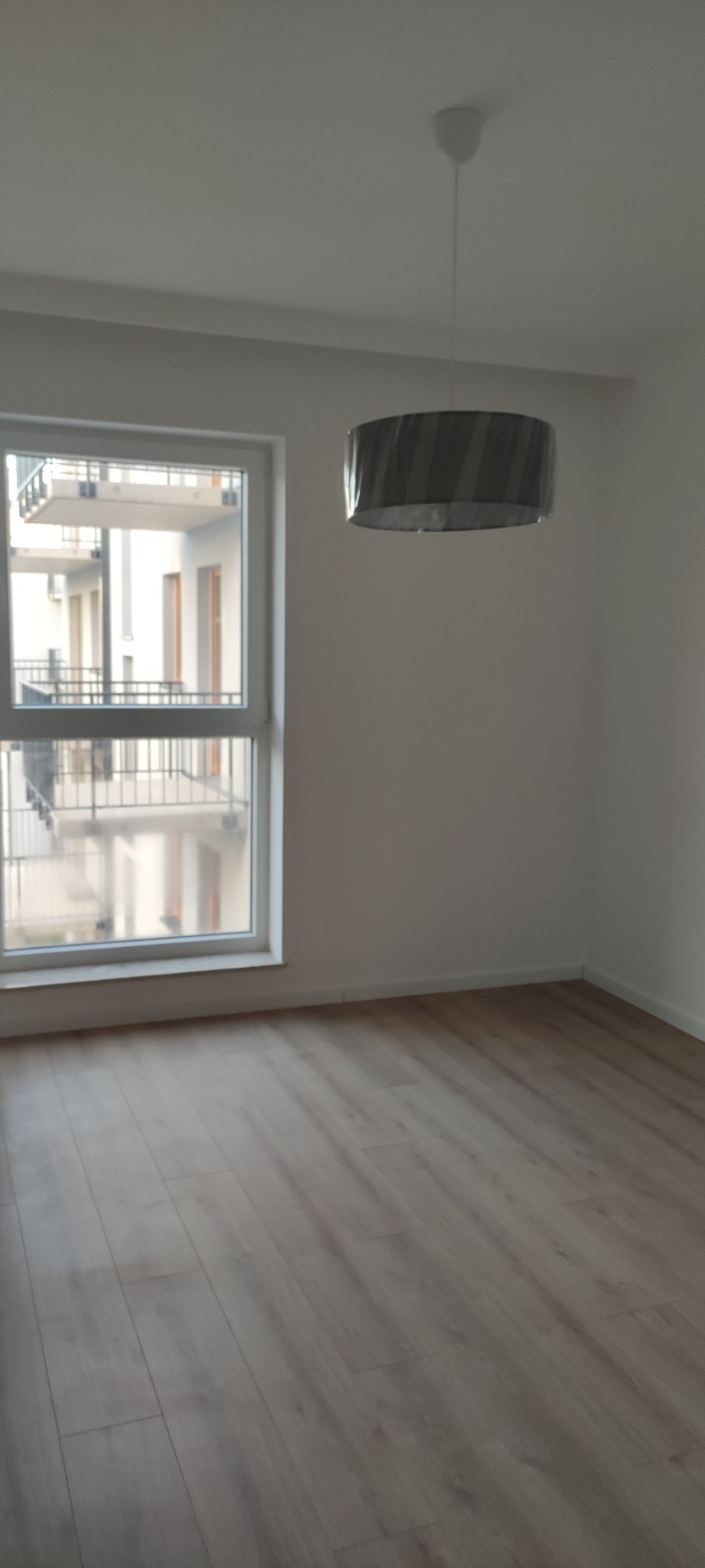 Pusty pokój z panelami podłogowymi w odcieniach szarości, widok na balkon z metalową barierką przez okno, szary abażur lampy wiszącej.