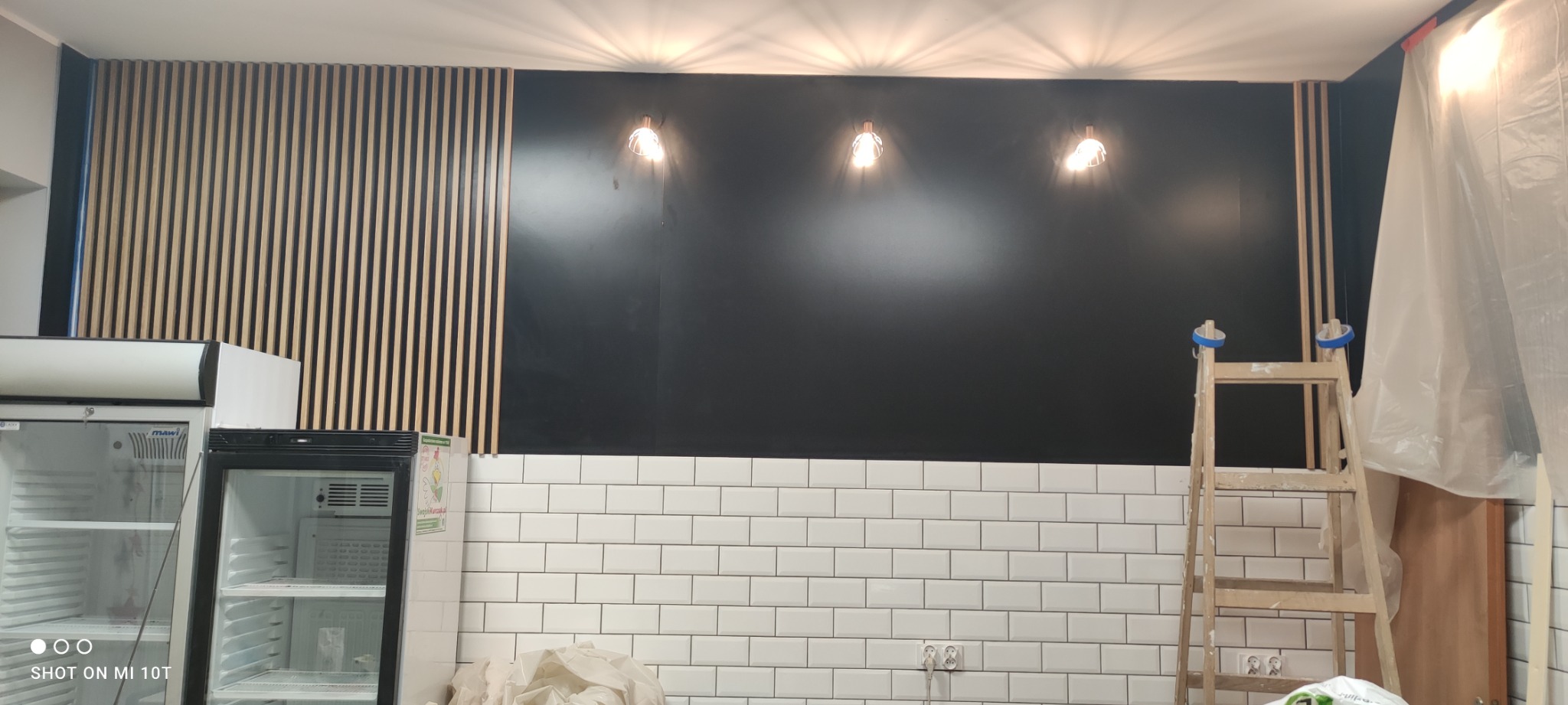 Wnętrze lokalu gastronomicznego w trakcie wykańczania: czarna ściana z trzema lampami, fragment ściany z drewnianymi lamelami, białe kafelki, drabina malarska pod ścianą, zakryta folią część...