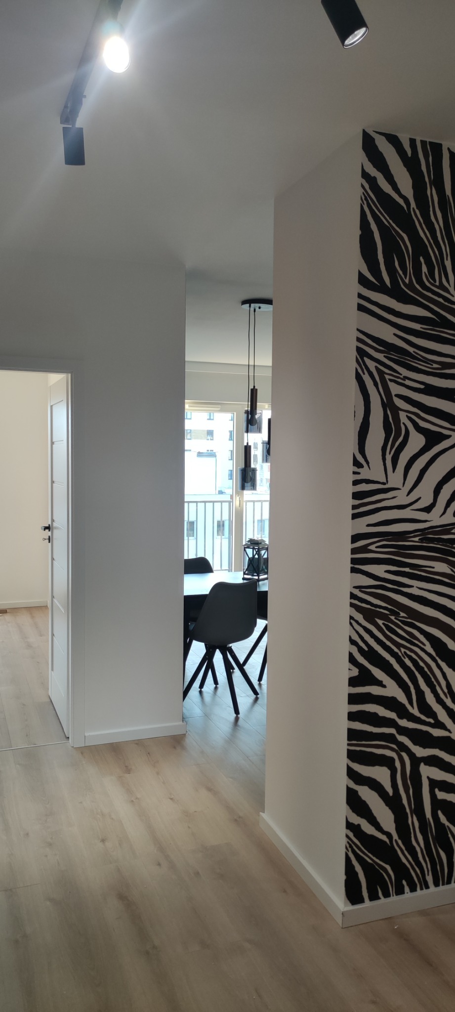 Nowoczesne wnętrze mieszkania z drewnianą podłogą, widokiem na stół z krzesłami, oświetleniem sufitowym i ścianą z motywem zebry.