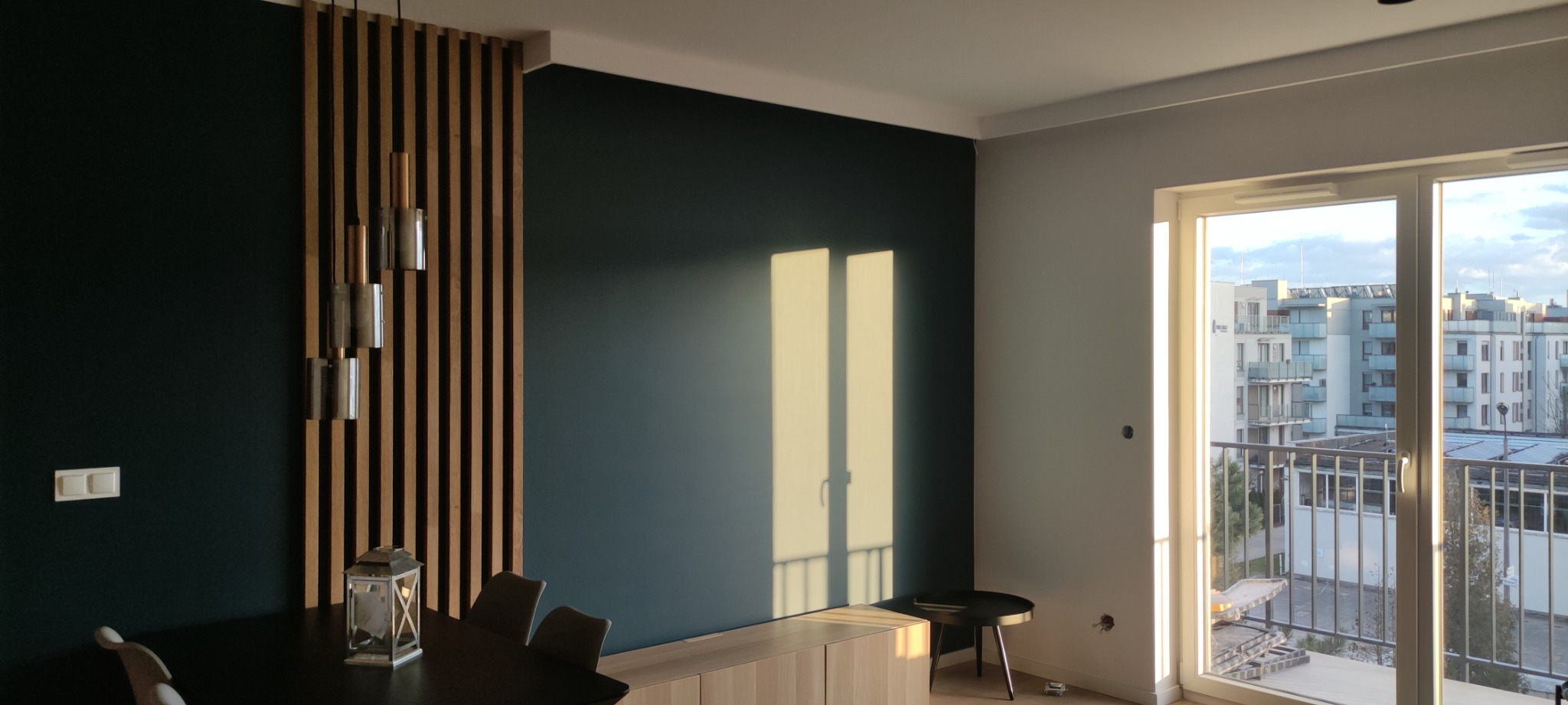 Nowoczesne wnętrze z ciemnozieloną ścianą, drewnianą ażurową ścianką działową, wiszącymi lampami i widokiem na osiedle przez balkonowe okno.