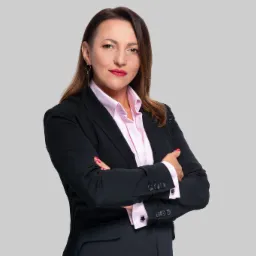 Portret kobiety w czarnym garniturze i różowej koszuli, z założonymi rękami, na jednolitym szarym tle.