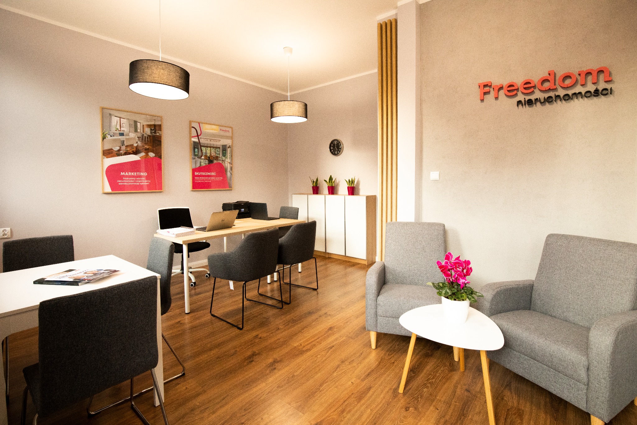Jasne biuro nieruchomości z drewnianą podłogą, szarymi fotelami, biurkami z laptopami i plakatami na ścianach. Logo 'Freedom nieruchomości' na ścianie.