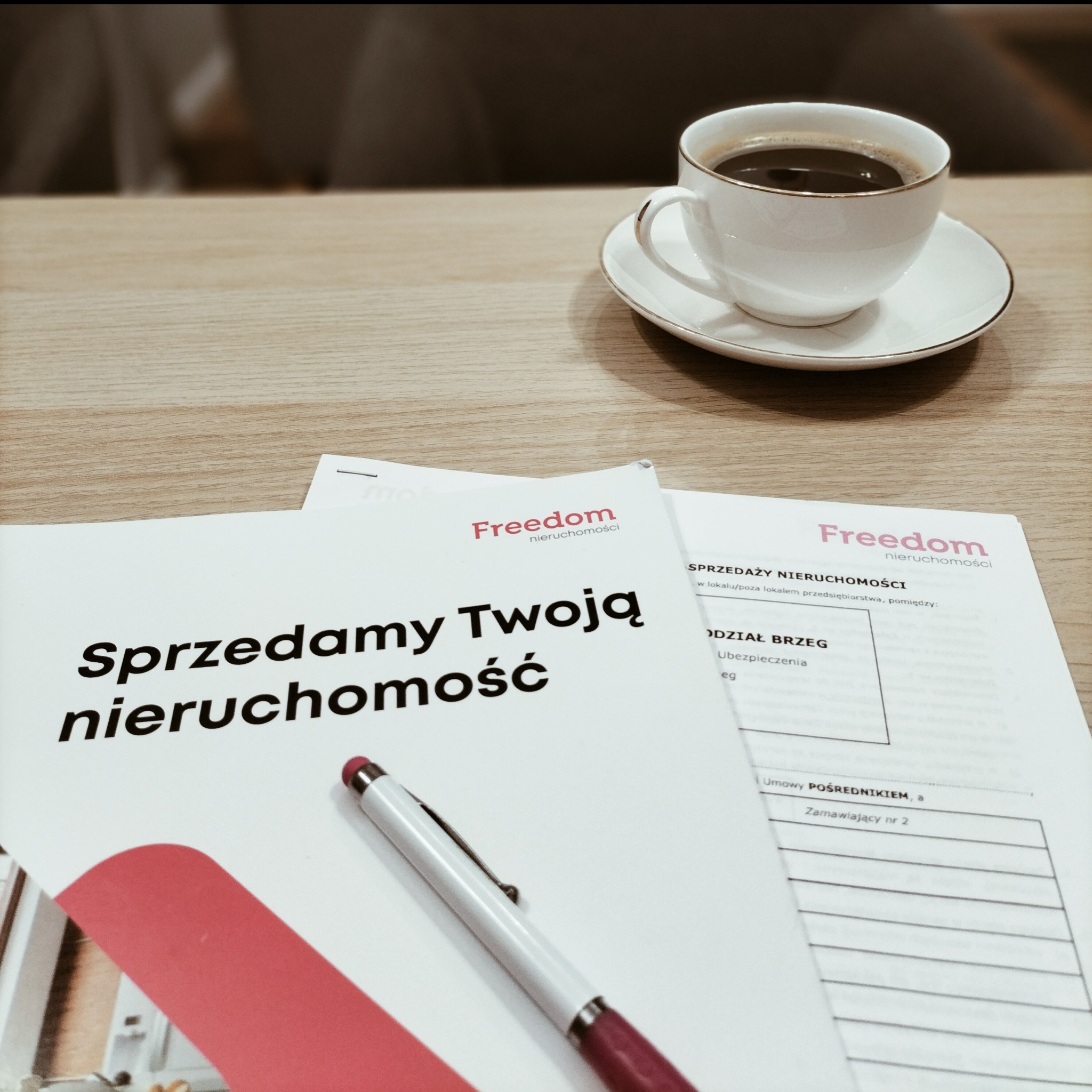 Kawa na biurku obok dokumentów z nagłówkiem 'Sprzedamy Twoją nieruchomość' i długopisu, z logo Freedom nieruchomości.