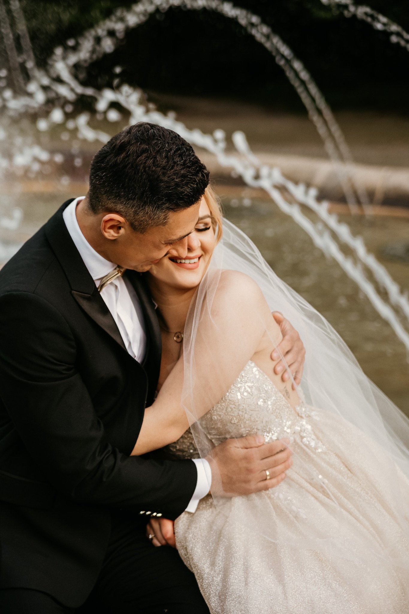 Uśmiechnięta panna młoda w sukni zdobionej cekinami, obejmowana przez pana młodego w czarnym smokingu z muszką w kolorze złota, na tle fontanny.