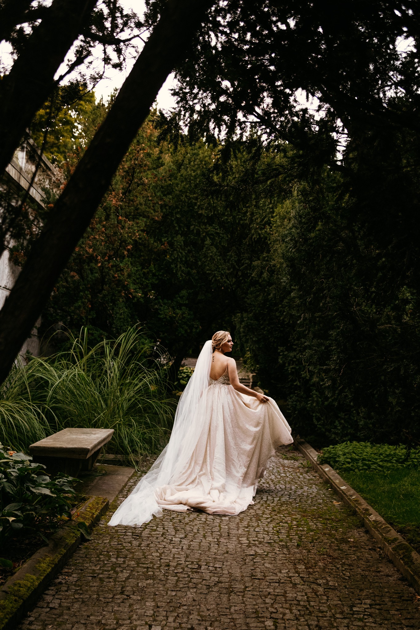 Panna młoda w sukni z odkrytymi plecami i długim welonem spaceruje po kamiennej ścieżce w ogrodzie, trzymając brzeg sukni. W tle gęsta zieleń i fragmenty architektury.