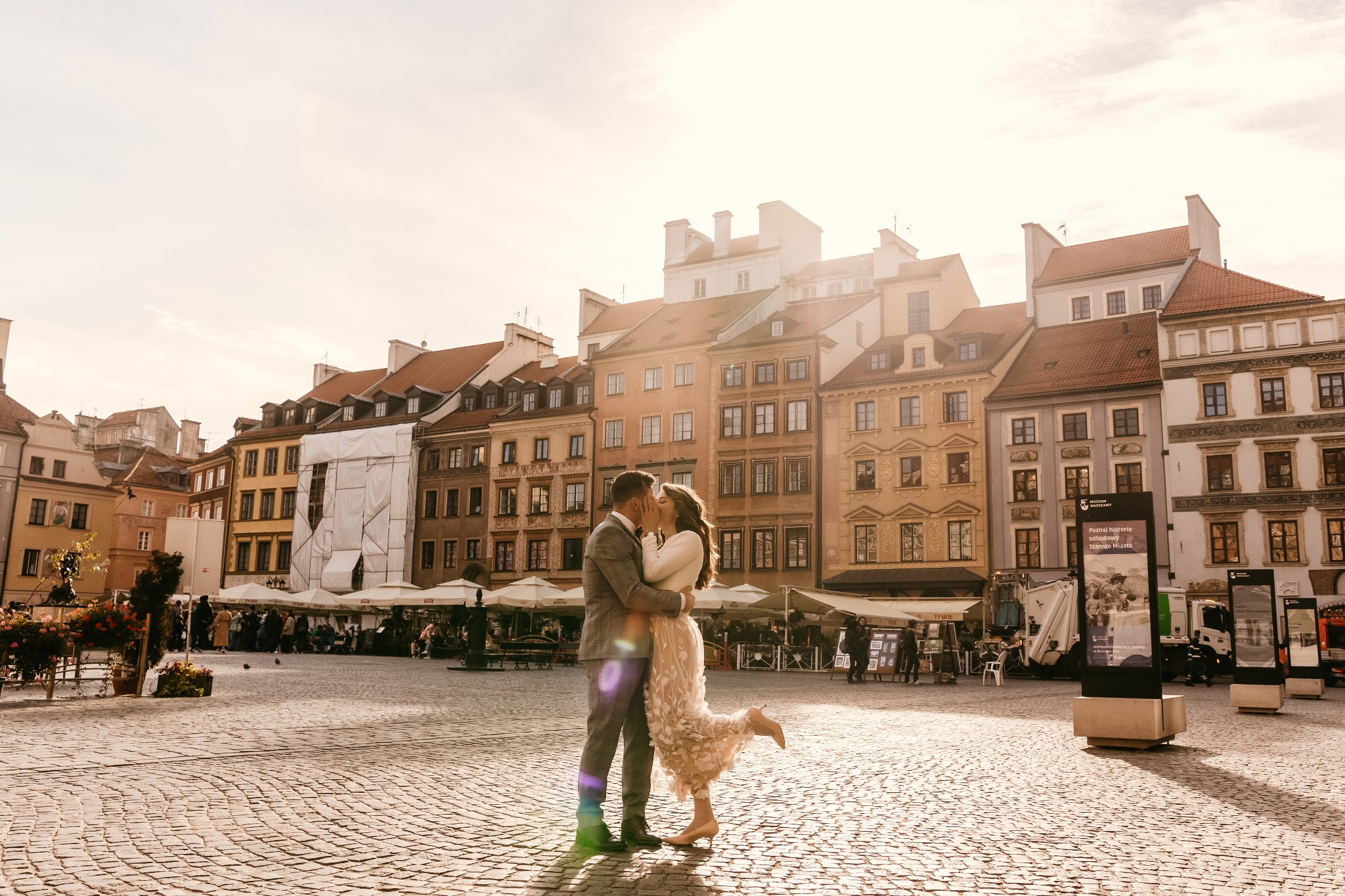 Para młoda całująca się na brukowanym placu Starego Miasta w Warszawie, ona w zwiewnej sukience z kwiatowymi aplikacjami i kremowych szpilkach, on w szarym garniturze, w tle zabytkowe kamienice...