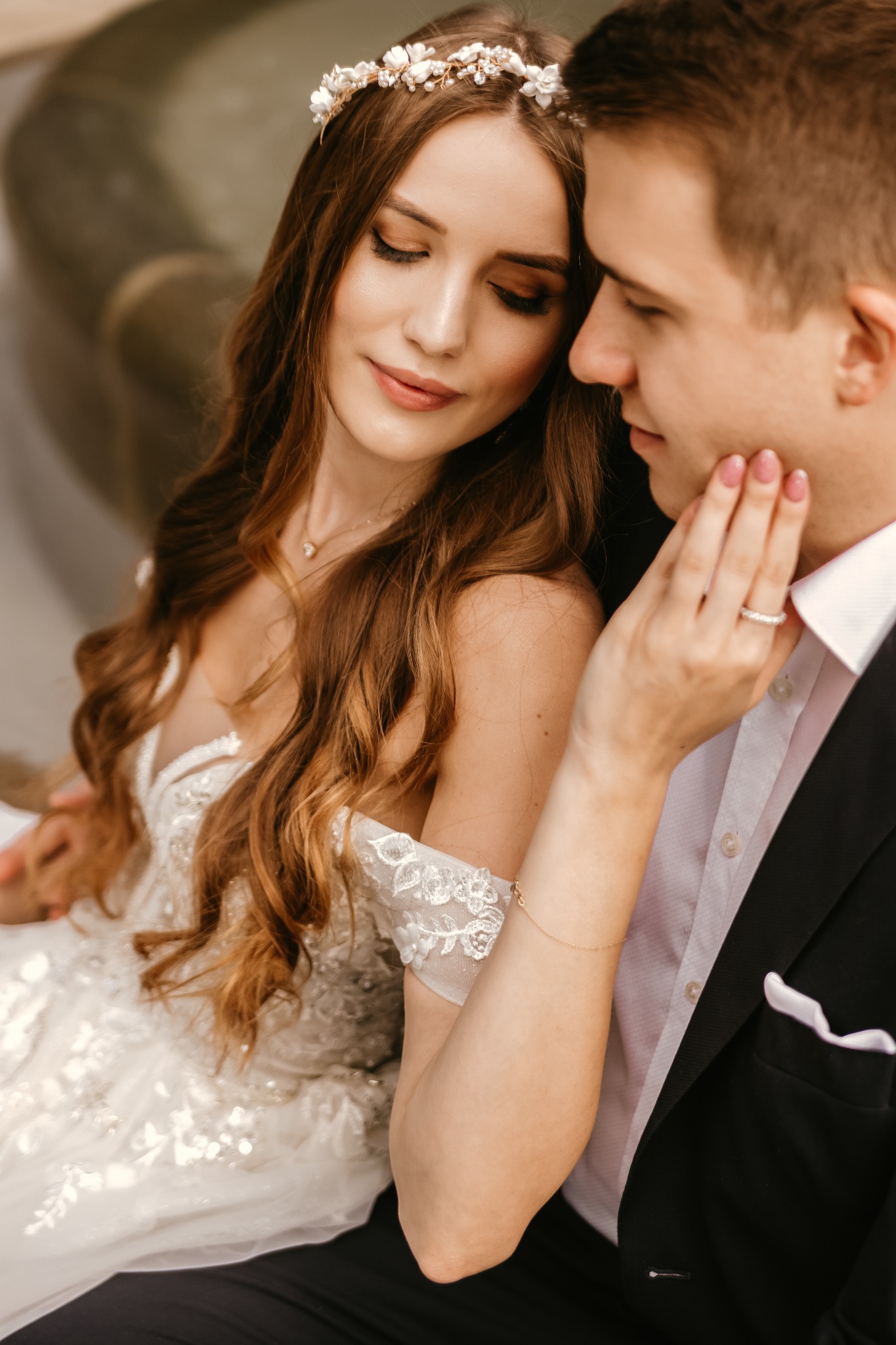 Uśmiechnięta panna młoda w koronkowej sukni i kwiecistej ozdobie na głowie, delikatnie dotykana przez pana młodego w czarnym garniturze, oboje zamyśleni i patrzący w dół.