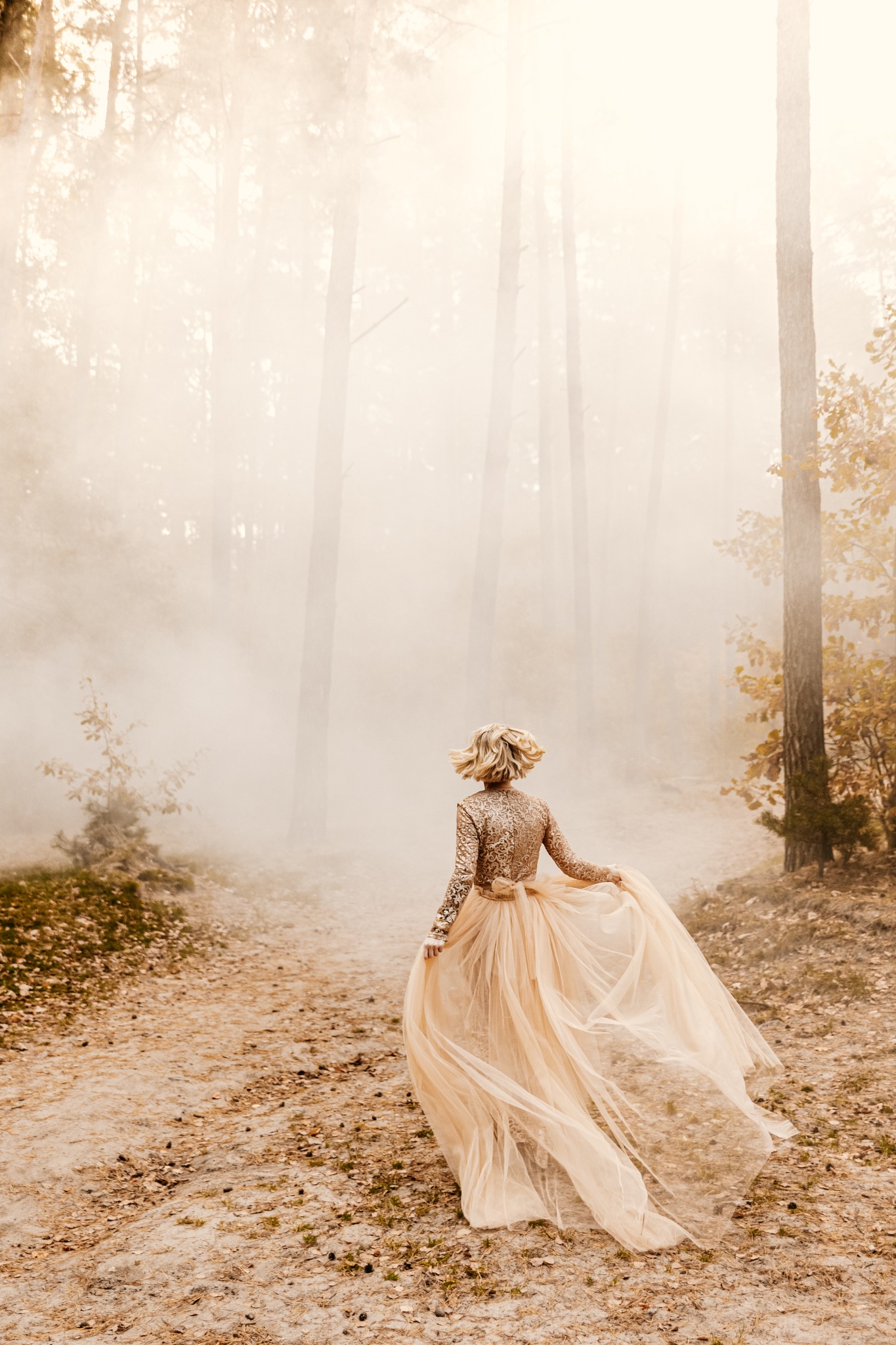 Kobieta w złotej sukni z tiulowym dołem biegnie ścieżką w lesie spowitym mgłą, tyłem do obiektywu.