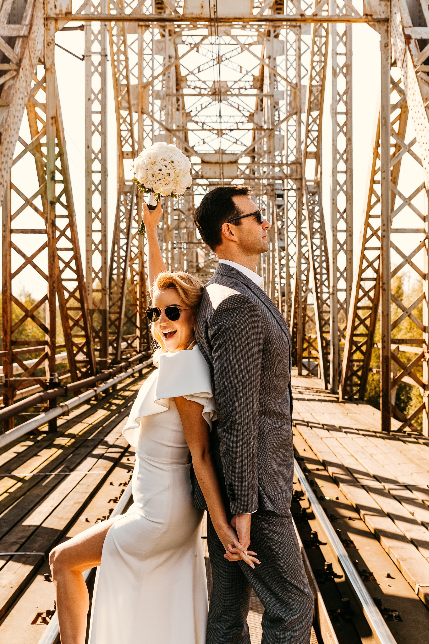 Uśmiechnięta panna młoda w okularach przeciwsłonecznych, trzymająca bukiet białych róż, opiera się plecami o pana młodego w szarym garniturze na tle konstrukcji stalowego mostu.