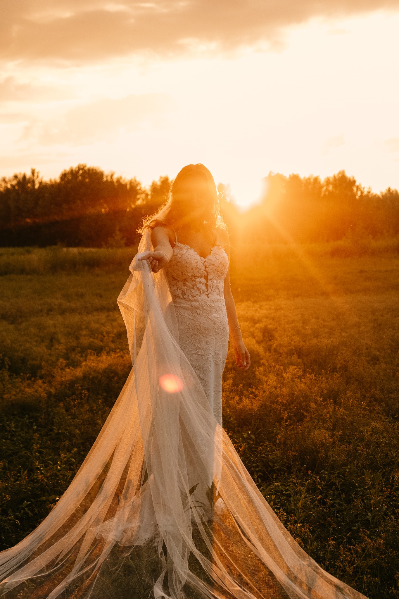 Panna młoda w koronkowej sukni ślubnej z długim welonem, stojąca na łące w promieniach zachodzącego słońca, wyciąga dłoń w stronę widza.