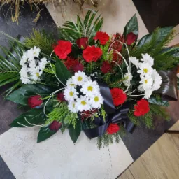 Kompozycja kwiatowa z czerwonych goździków, białych margaretek, czerwonych róż, zielonych liści i czarnej wstążki na podłodze w szachownicę.