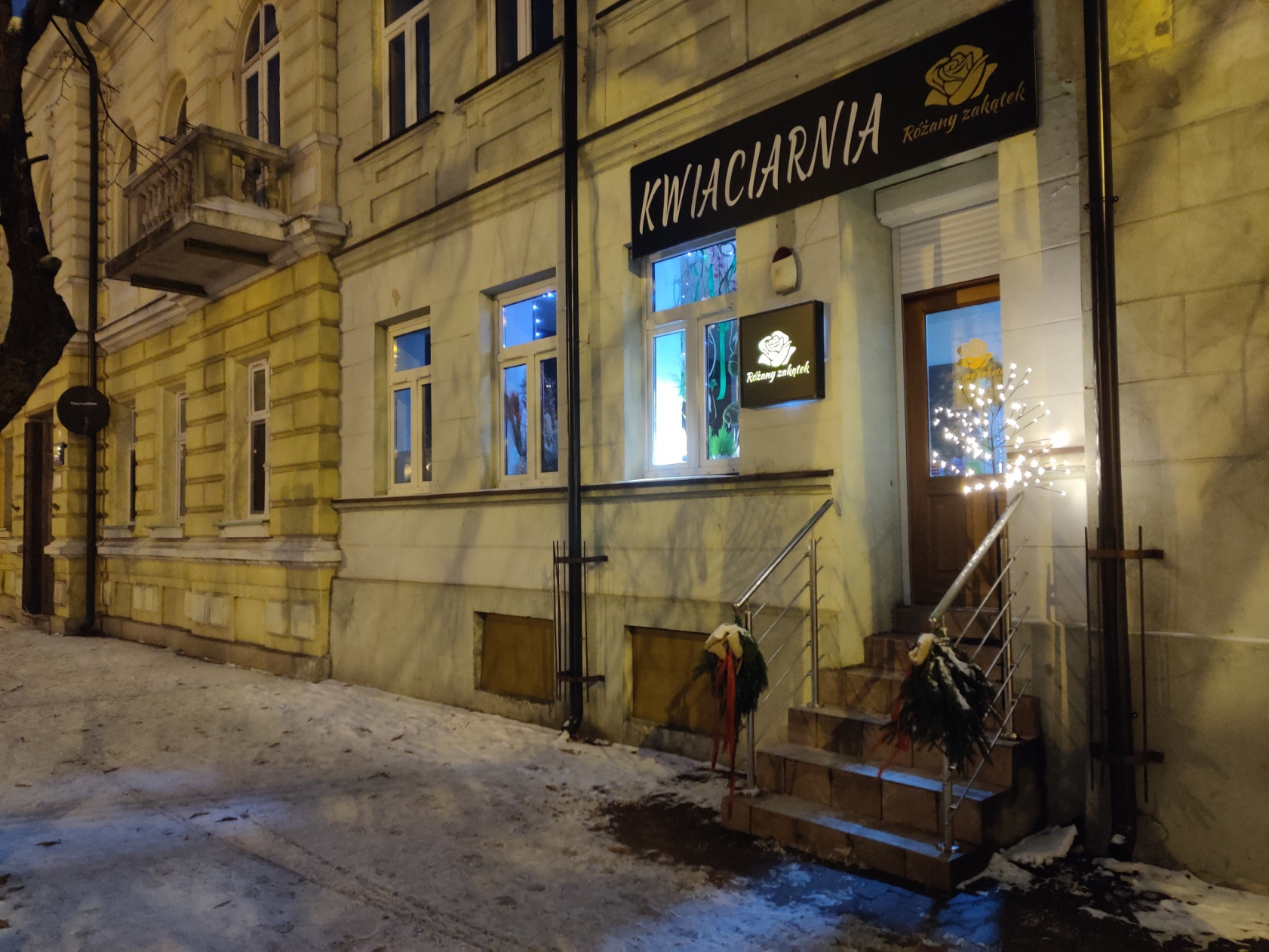 Zimowy wieczór przed kwiaciarnią 'Różany zakątek' z dekoracjami świątecznymi na schodach i podświetlanymi szyldami.