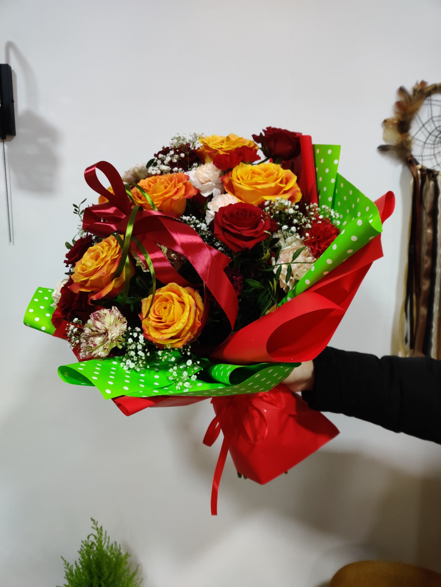 Bukiet mieszany z róż w odcieniach czerwieni, pomarańczy i żółci, owinięty w czerwony i zielony papier w białe kropki, przewiązany czerwoną wstążką, trzymany w dłoni na tle białej ściany z łapaczem...