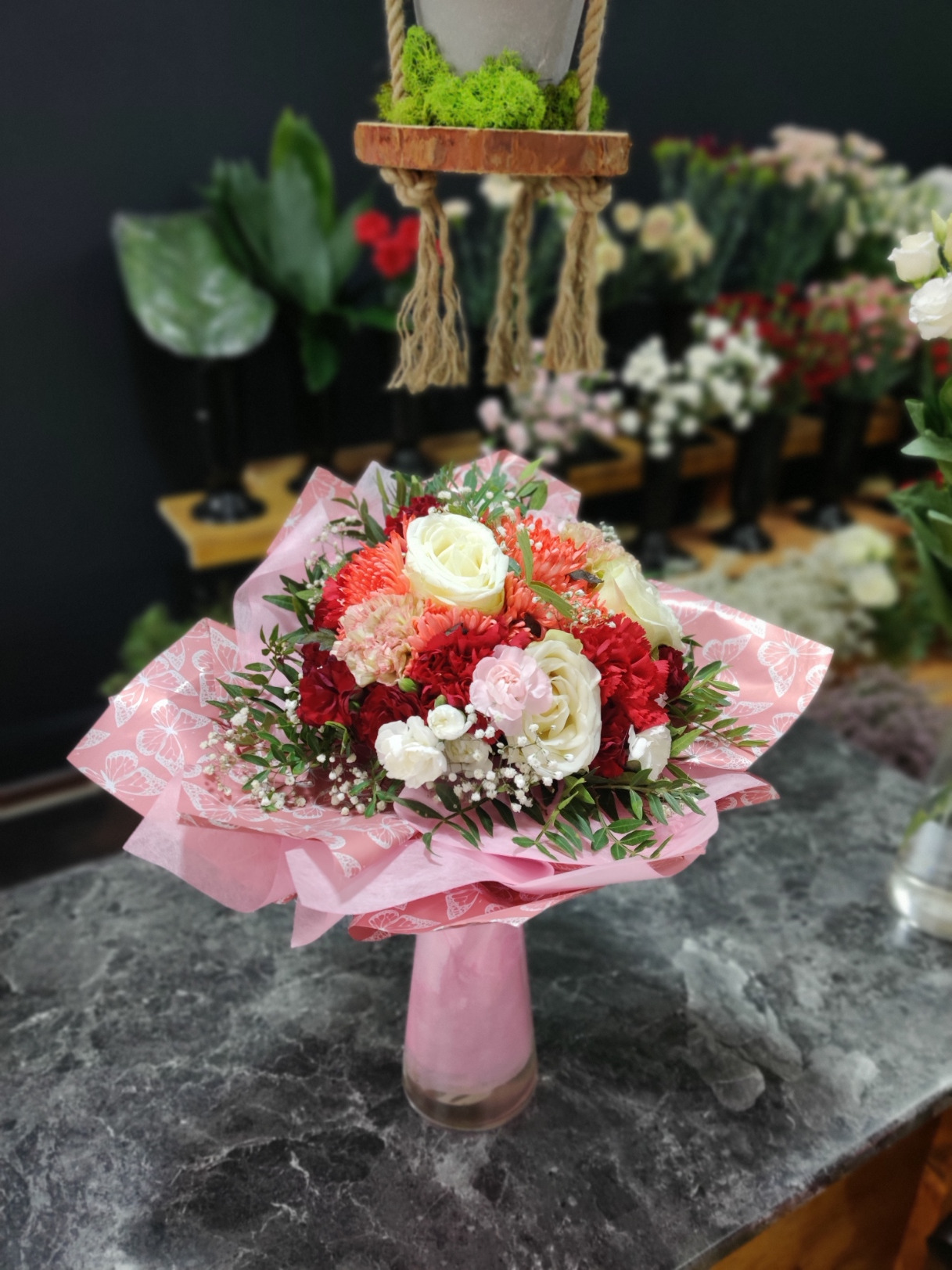 Elegancki bukiet z czerwonych i białych goździków oraz róż, zapakowany w różowy papier z motywem motyli, umieszczony w różowym wazonie na marmurowym blacie. W tle widoczne inne kwiaty i dekoracje.