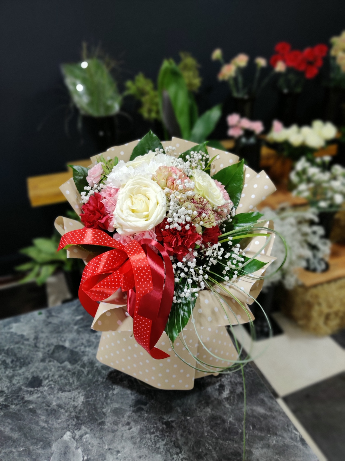 Ułożony bukiet z białych róż, czerwonych i różowych goździków, oraz gipsówki, owinięty w papier w kropki i przewiązany czerwoną wstążką w białe kropki, na blacie z szarego marmuru.