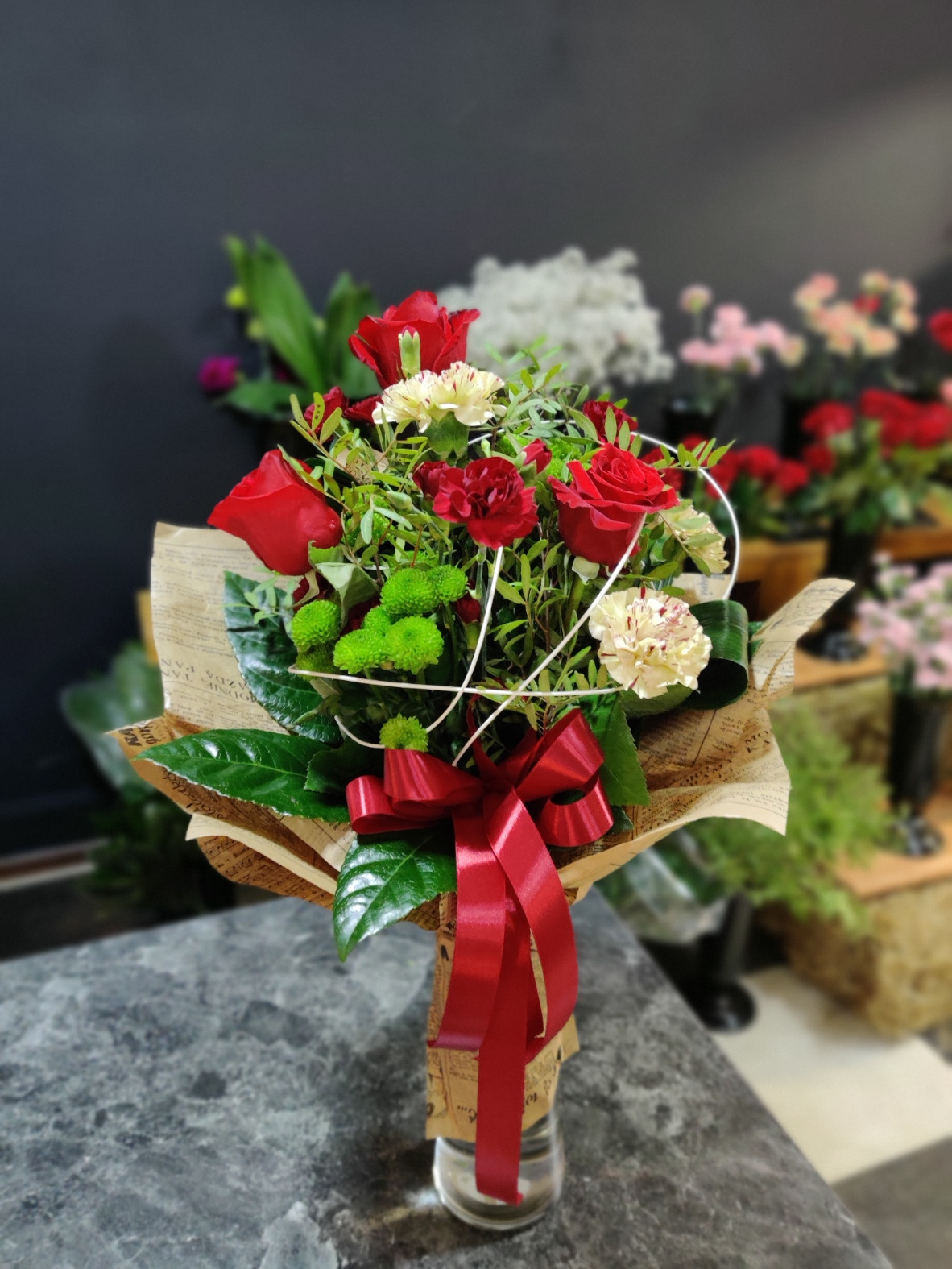 Bukiet czerwonych róż, goździków i zielonych chryzantem, przewiązany czerwoną wstążką, zapakowany w papier z nadrukiem w stylu retro, umieszczony w szklanym wazonie, widoczny na tle kwiaciarni...