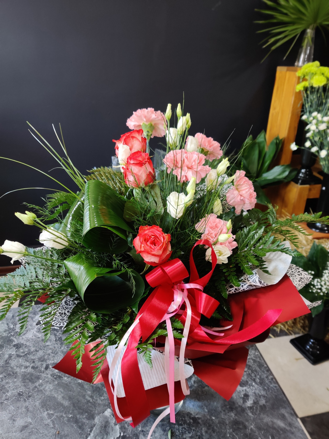 Elegancki bukiet mieszany z róż, goździków i eustomy, przewiązany czerwoną wstążką, na tle ciemnej ściany i fragmentu drewnianej konstrukcji z zielonymi roślinami.