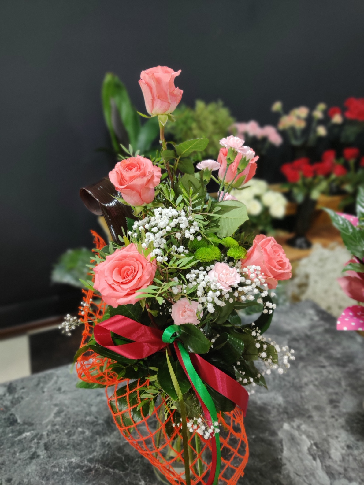 Dekoracyjny bukiet z różowych róż, białych gipsówek, zielonych liści i czerwono-zielonej wstążki, umieszczony w pomarańczowej siatce, na tle rozmytych kwiatów.