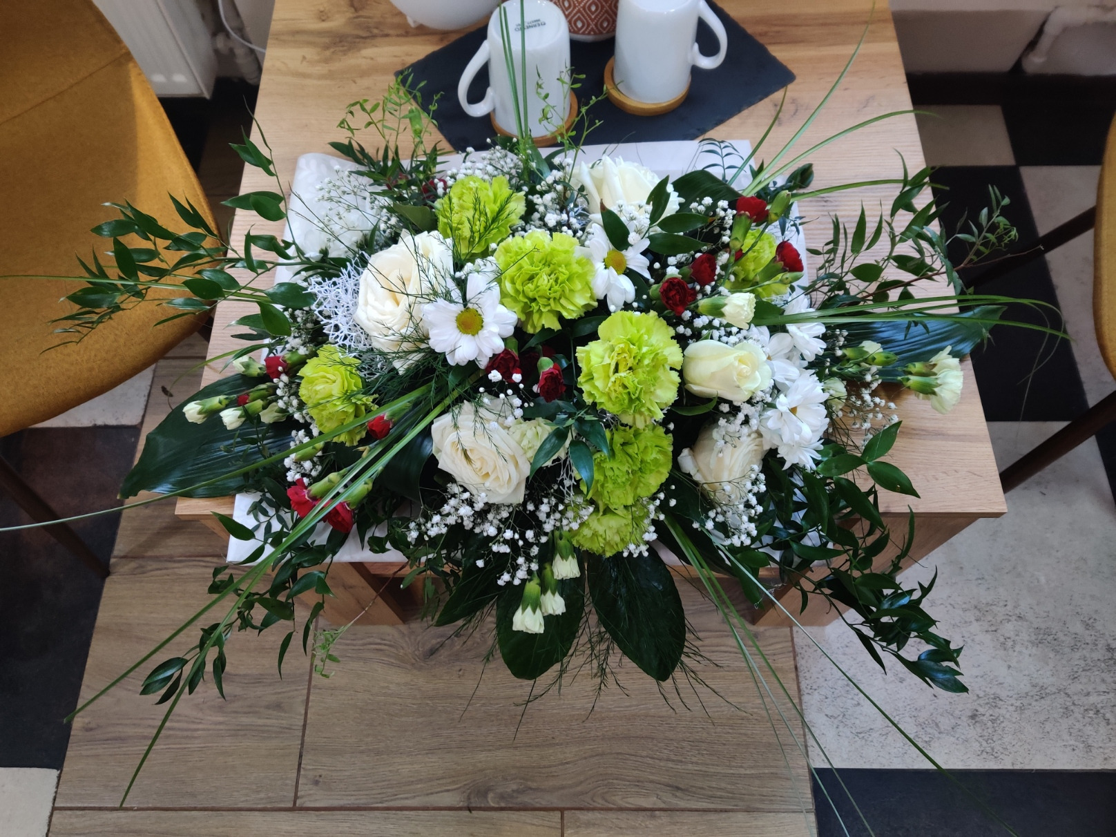 Kompozycja kwiatowa z białych róż, zielonych goździków i czerwonych akcentów na drewnianym stole, widok z góry, obok dwie białe filiżanki.