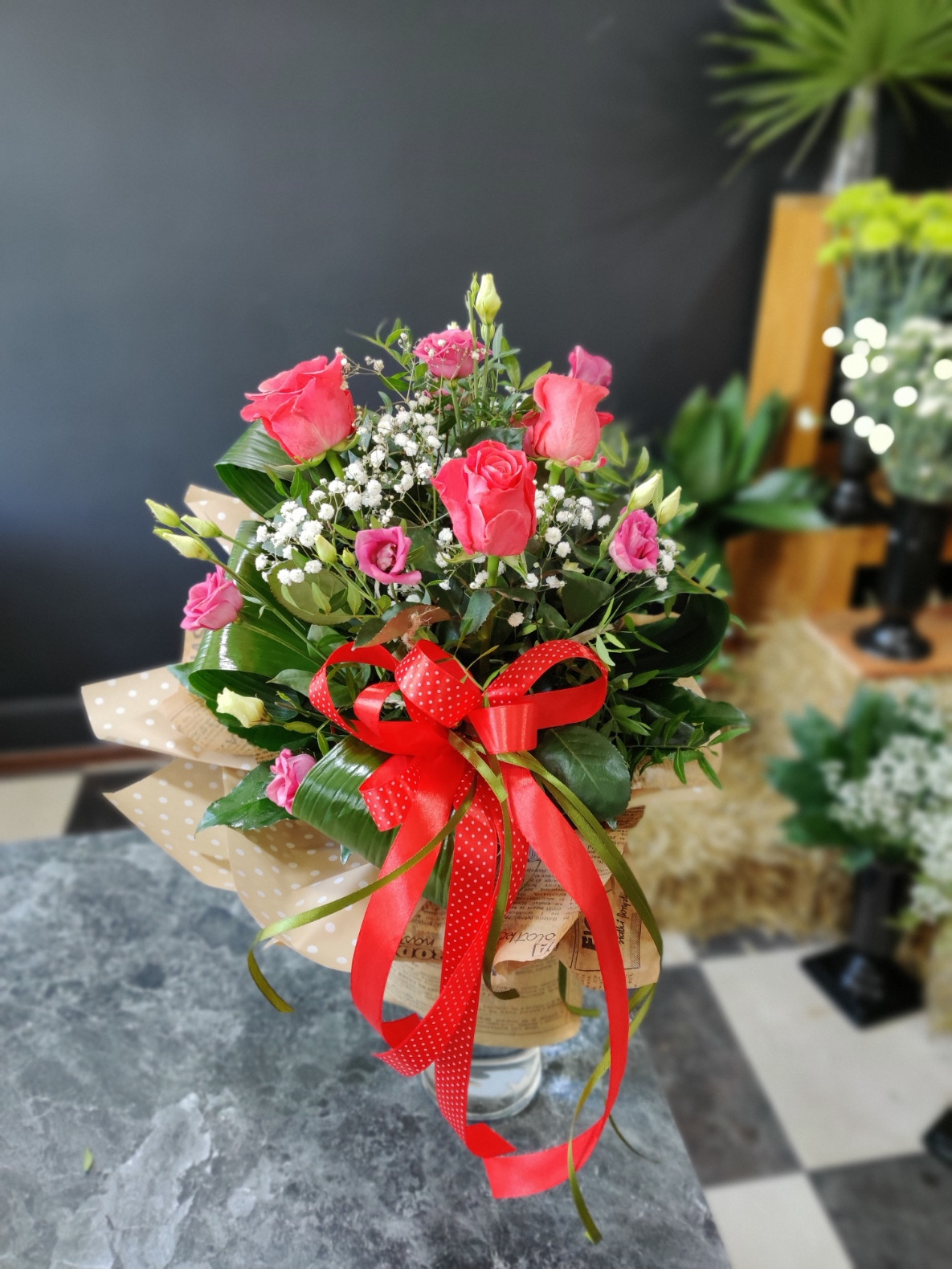 Kompozycja kwiatowa z różowych róż, eustomy i gipsówki, związana czerwoną wstążką w białe kropki, zapakowana w papier z nadrukiem imitującym gazety, na tle ciemnej ściany i rozmytych roślin.
