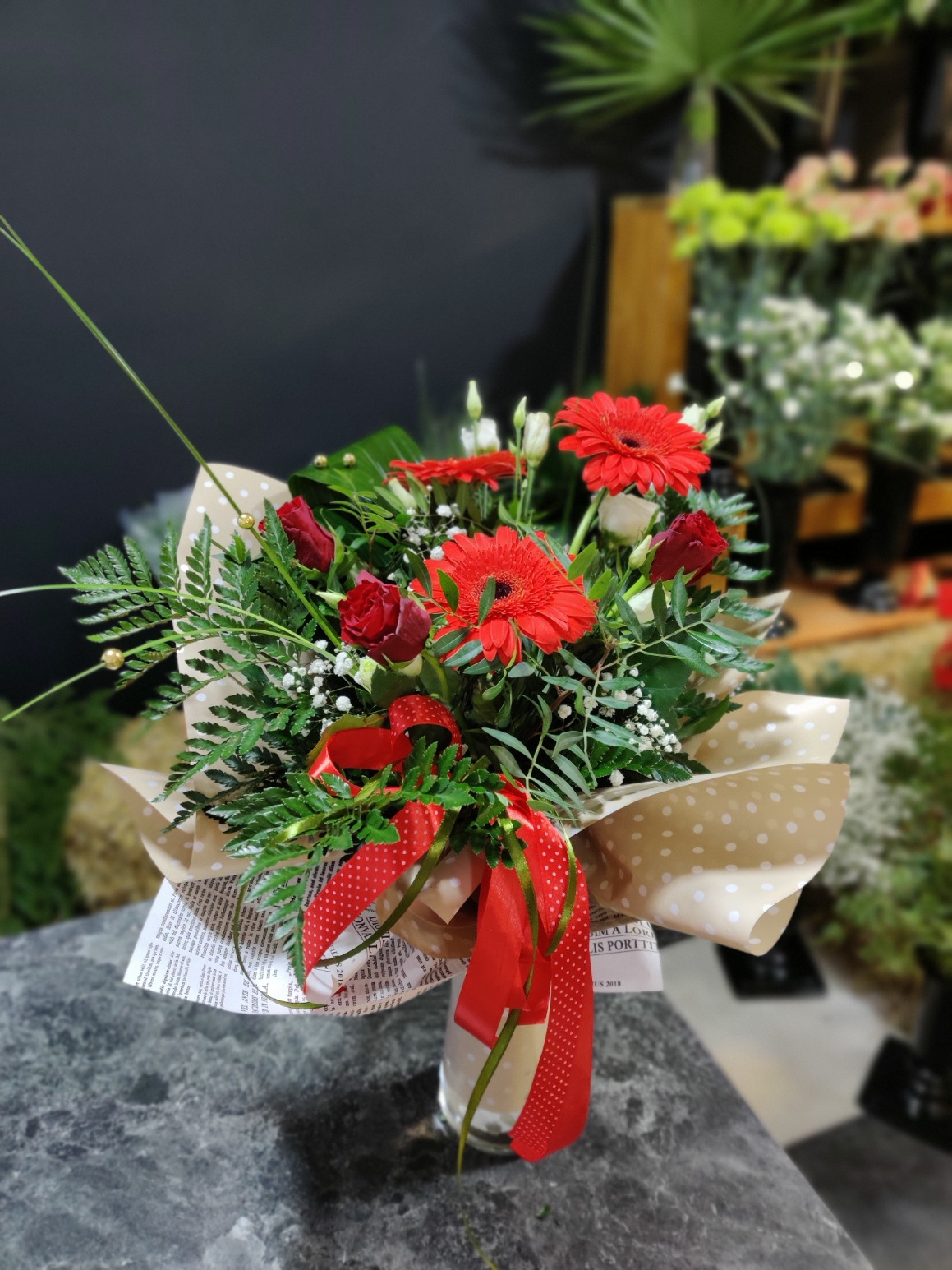 Kompozycja kwiatowa z czerwonych gerber, róż i zielonych paproci, owinięta w papier w kropki i przewiązana czerwoną wstążką w kropki, umieszczona w szklanym wazonie na szarym blacie, w tle rozmyte...