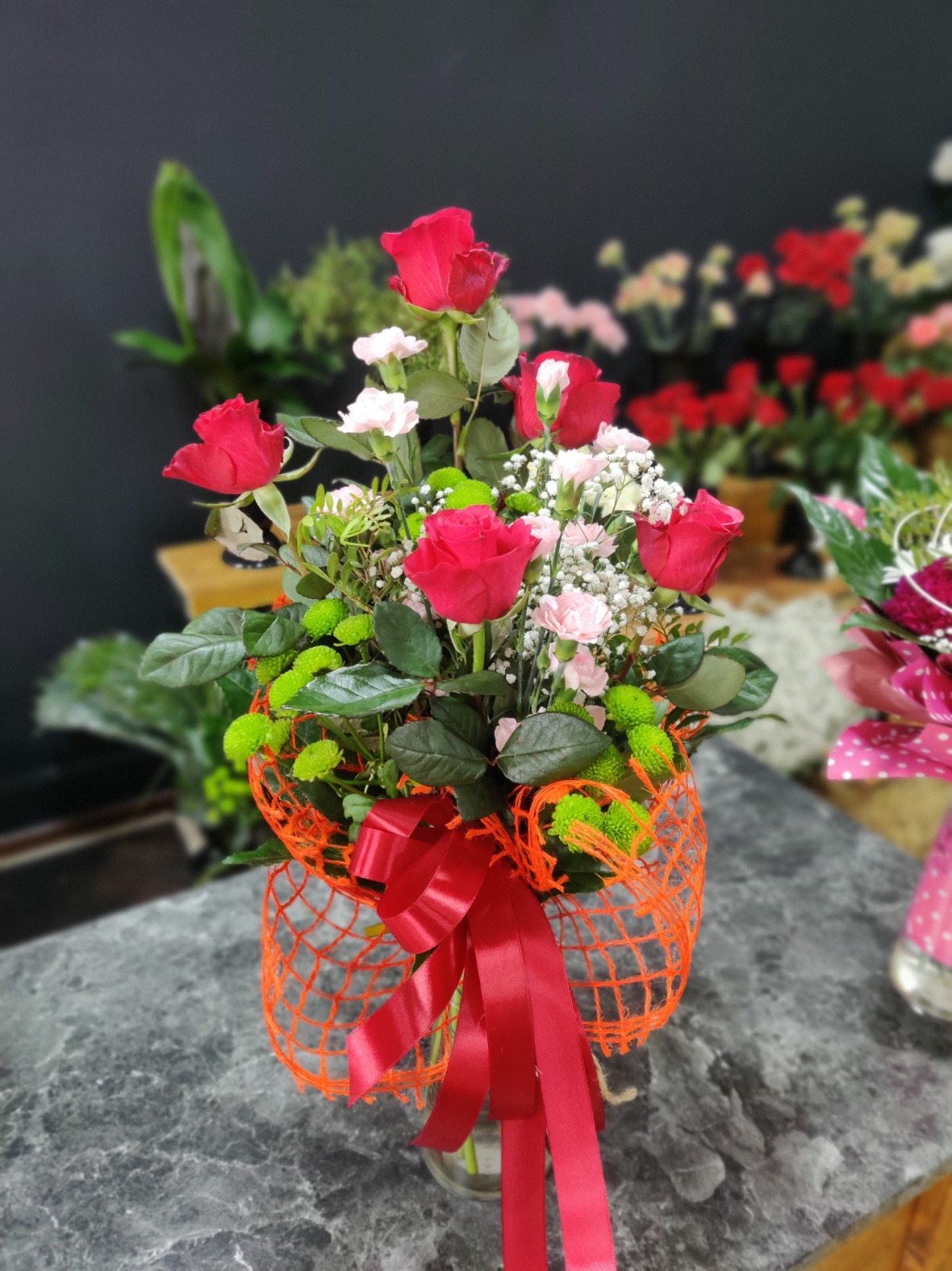 Bujny bukiet z czerwonych róż, białych goździków i zielonych chryzantem, ozdobiony pomarańczową siatką i czerwoną satynową wstążką, na tle ciemnego wnętrza kwiaciarni.