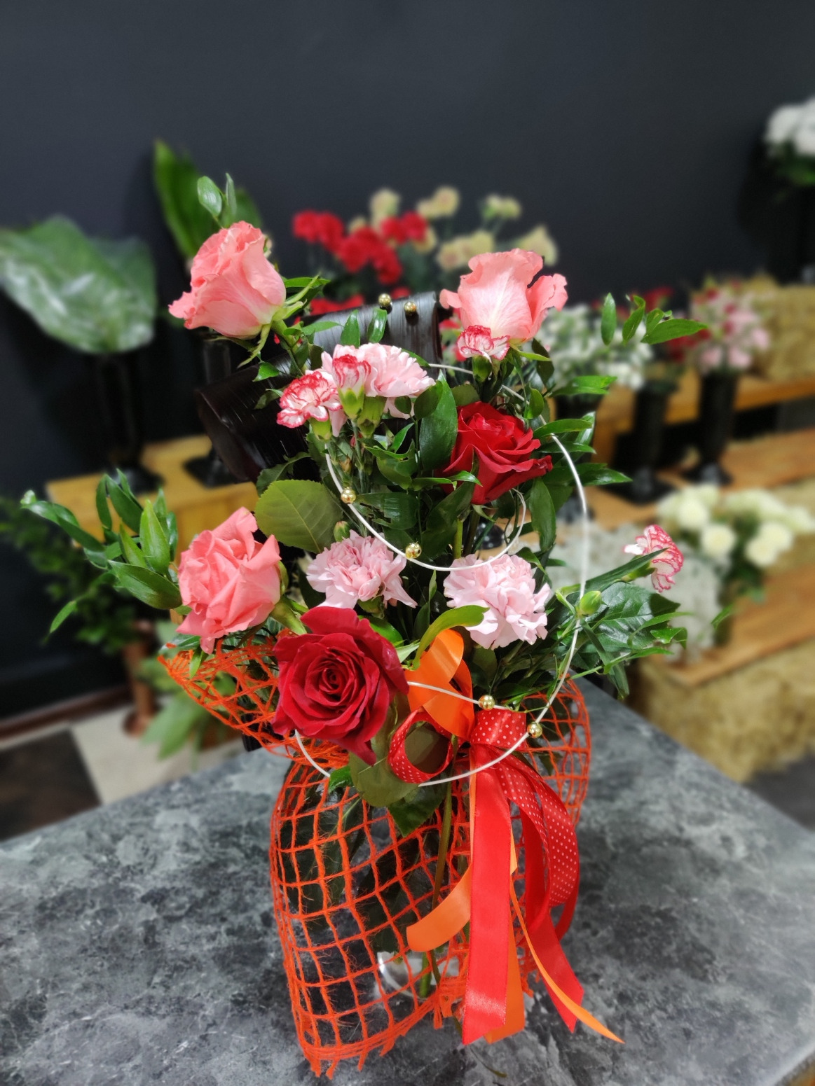 Dekoracyjny bukiet z czerwonych i różowych róż, różowych goździków, zieleni oraz pomarańczowej siatki i czerwonej wstążki w kropki, ustawiony na szarym blacie.