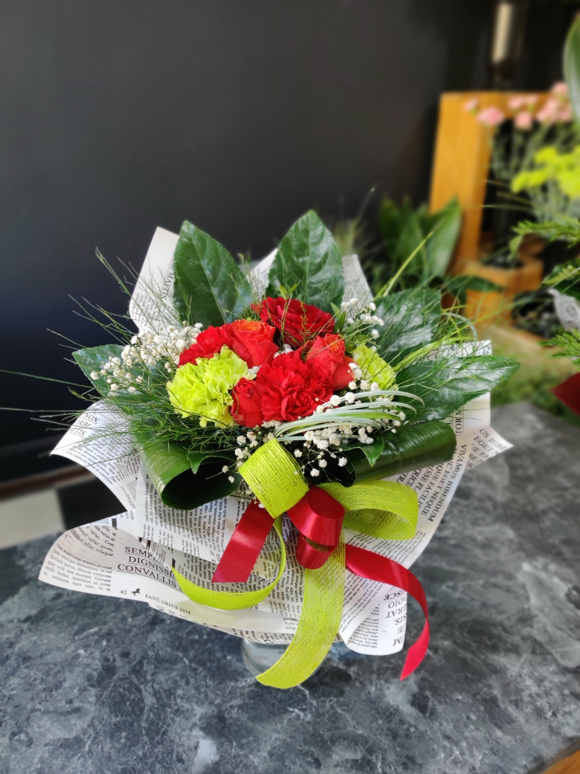 Kompozycja kwiatowa z czerwonych i limonkowych goździków oraz białych drobnych kwiatów, owinięta papierem z nadrukiem tekstu i przewiązana wstążkami w kolorach czerwonym i limonkowym. Widoczne...