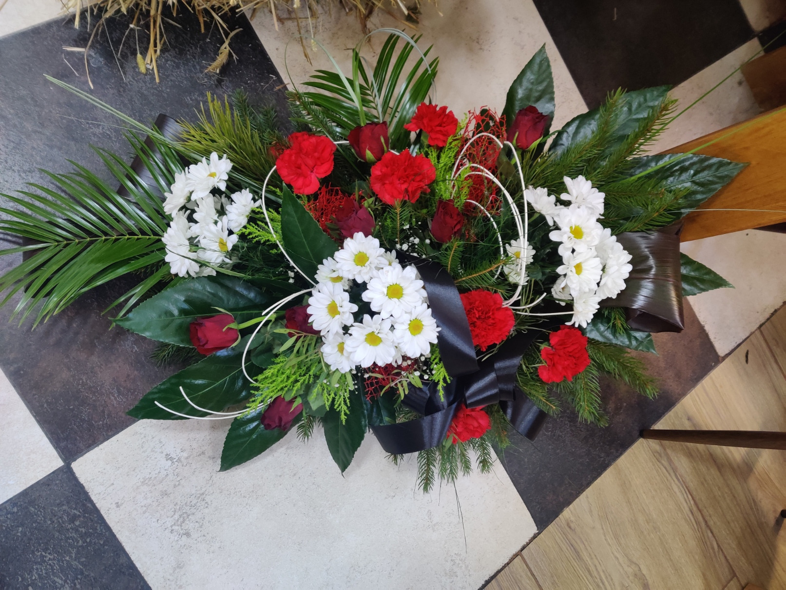 Kompozycja kwiatowa z czerwonych goździków, białych margaretek, czerwonych róż, zielonych liści i czarnej wstążki na podłodze w szachownicę.