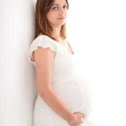 Portret kobiety w ciąży w białej sukience, opierającej się o białą ścianę, z delikatnym uśmiechem i dłonią na brzuchu.